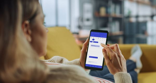 Junge Frau auf dem Sofa, in der Hand ihr Smartphone mit geöffneter Kalender-App.