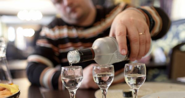 Gedeckter Tisch, im Vordergrund Vodkagläser,die von einem im unscharfen Hintergrund sitzenden Mann vollgeschenkt werden.