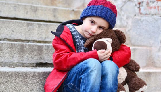 Kindergartenkind, ca. 5 Jahre alt, Mütze, Anorak, Jeans, einen Teddy im Arm, sitzt traurig auf einer grauen Steintreppe
