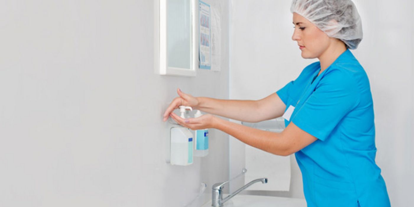Jüngere Frau in blauer OP-Arbeitskleidung mit Schutzhäubchen desinfiziert sich die Hände