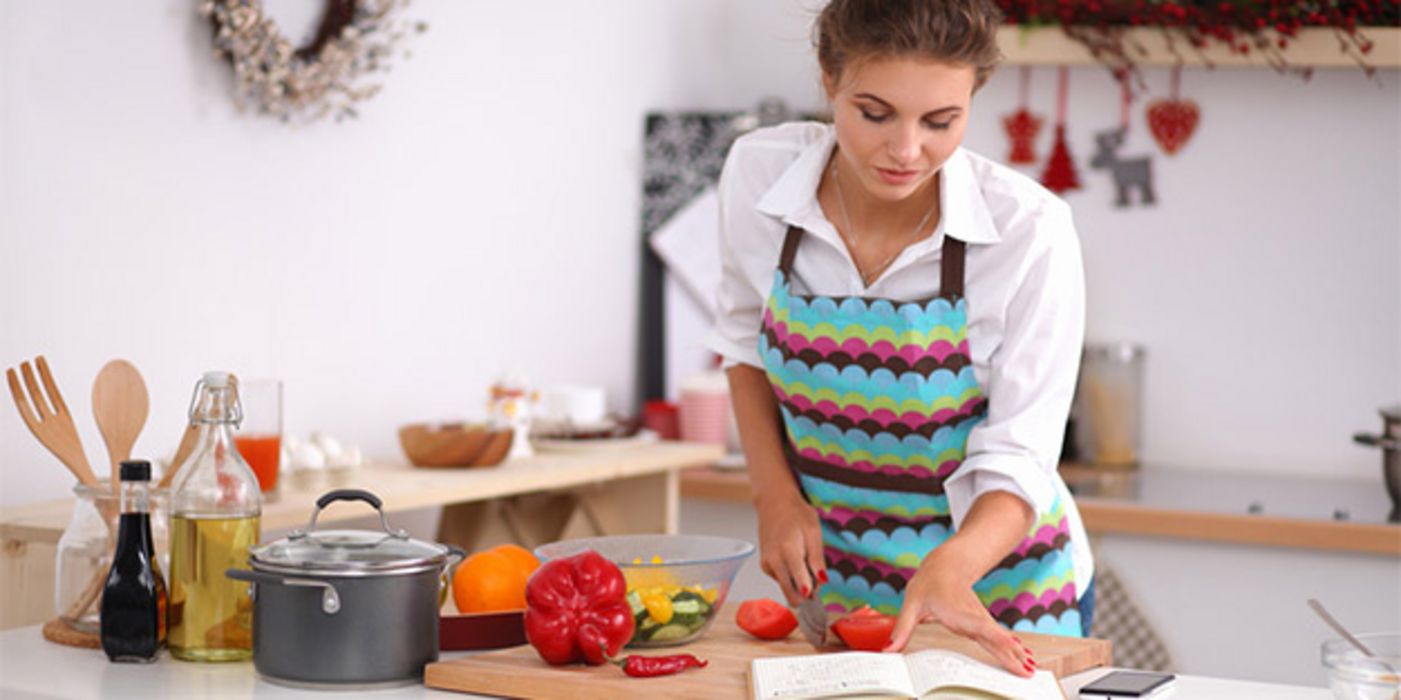 Frau in den 20ern mit bunter Schürze beim Kochen in Kochbuch blätternd