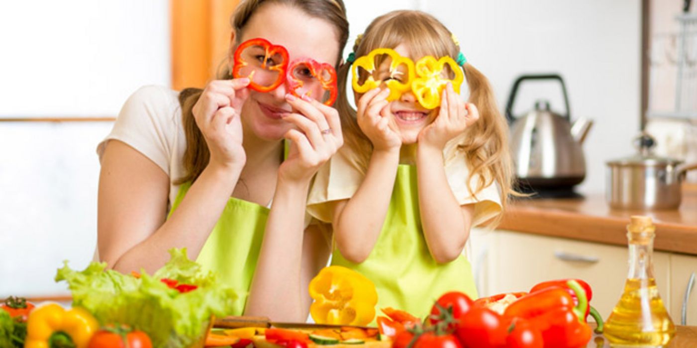 Mutter mit Kind, zusammen in der Küche, halten sich zwei Paprika-Scheiben vor die Augen.