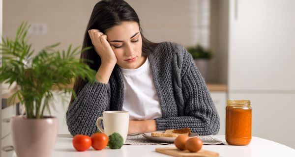 Junge Frau, sitzt vor einem Tisch mit Brot, Eiern und Gemüse und schaut verzweifelt nach unten.