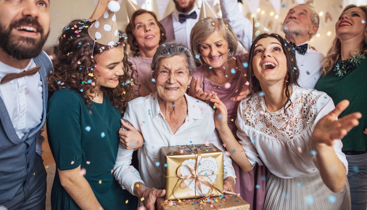 Eine ältere Dame hält Geschenke im Kreise ihrer Familie. Sie tragen Partyhüte und Konfetti fliegt durch die Luft.