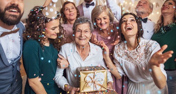 Eine ältere Dame hält Geschenke im Kreise ihrer Familie. Sie tragen Partyhüte und Konfetti fliegt durch die Luft.