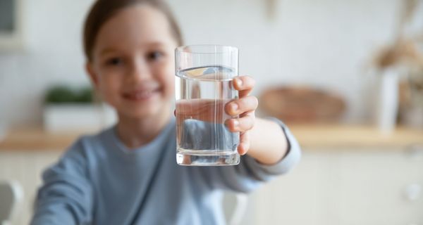 Mädchen, ca. 7 Jahre alt, streckt ein Glas mit Wasser in die Kamera, lächelt.