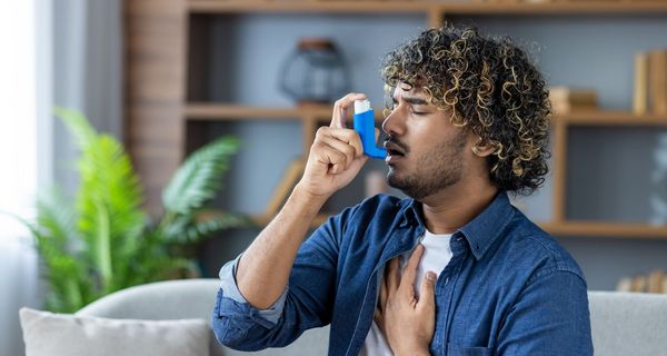 Junger Mann nutzt einen Inhalator. Er sitzt auf einem Sofa in einem Wohnzimmer.