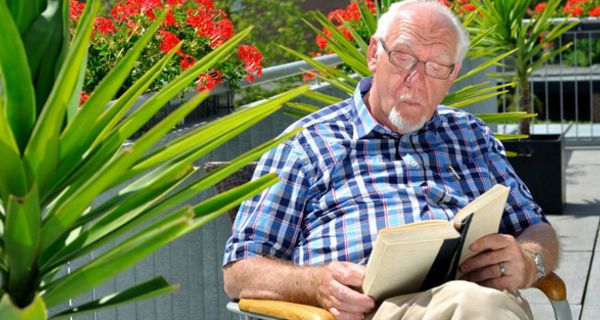 Senior sitzt auf sommerlichem Balkon und liest ein Buch