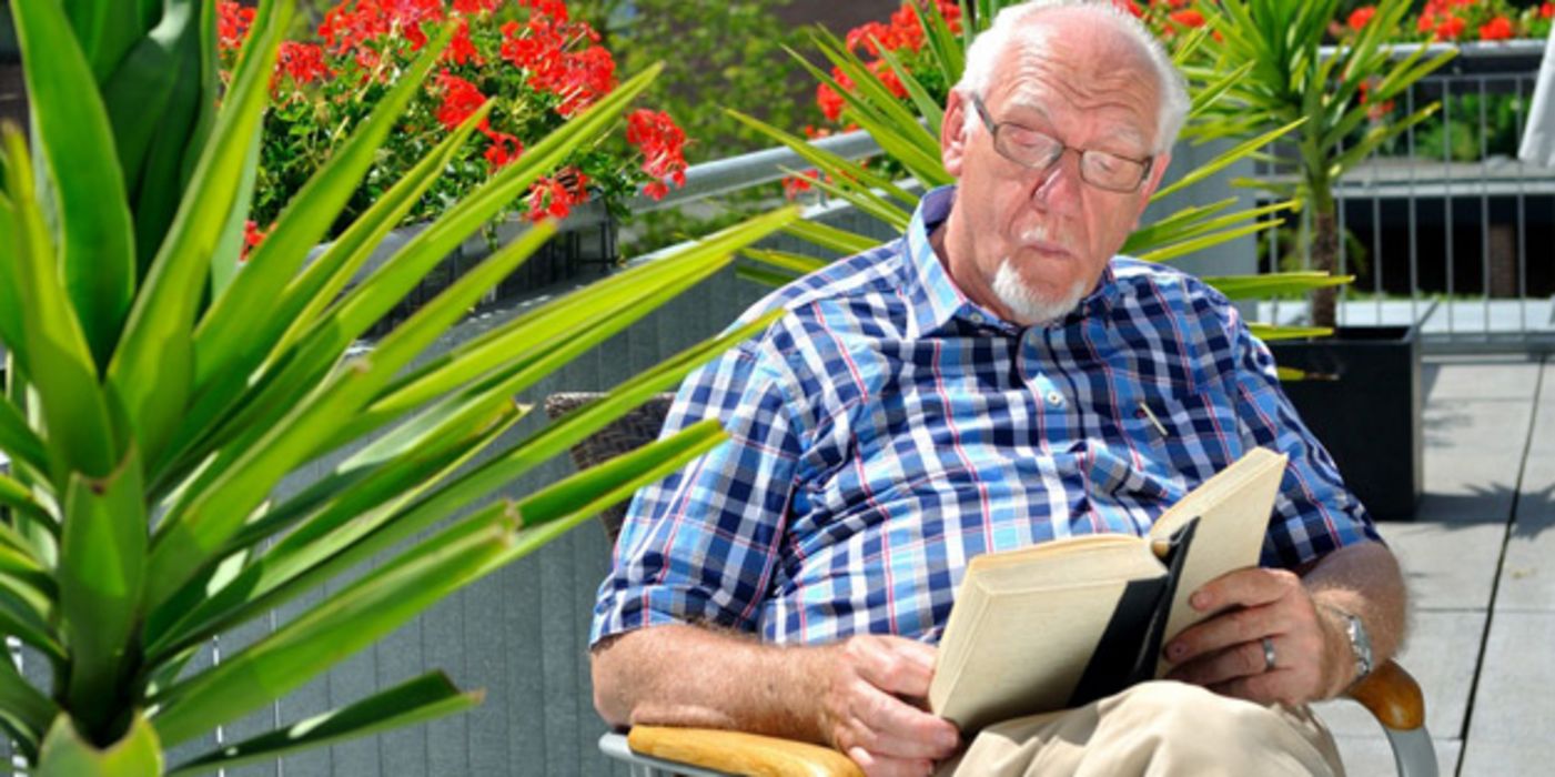Senior sitzt auf sommerlichem Balkon und liest ein Buch
