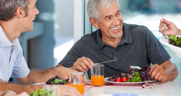 Zwei attraktive Männer über 60, grauhaarig, lachend, am Tisch mit Salatteller und Saft