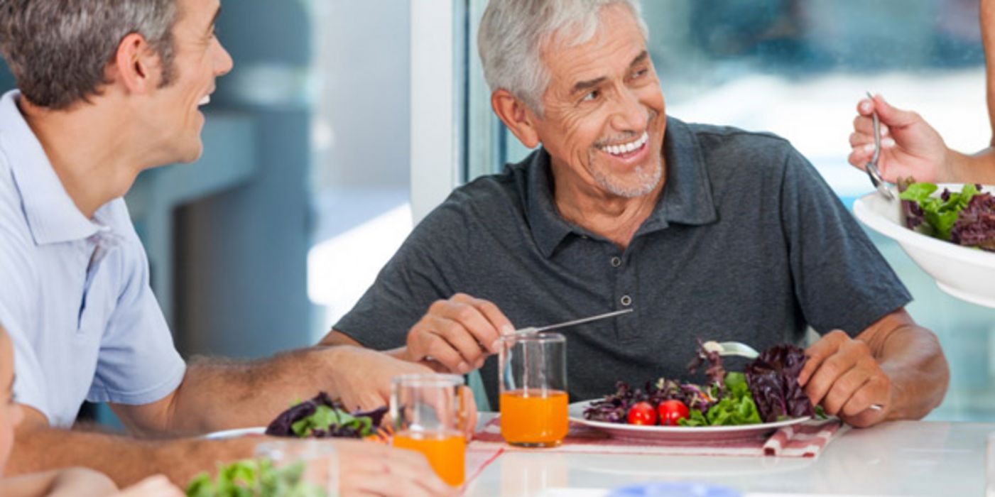 Zwei attraktive Männer über 60, grauhaarig, lachend, am Tisch mit Salatteller und Saft