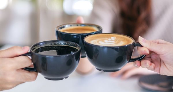 Drei Kaffeetassen in den Händen von Menschen. Im Hintergrund sind lange Haare und ein helles Oberteil zu erkenen.