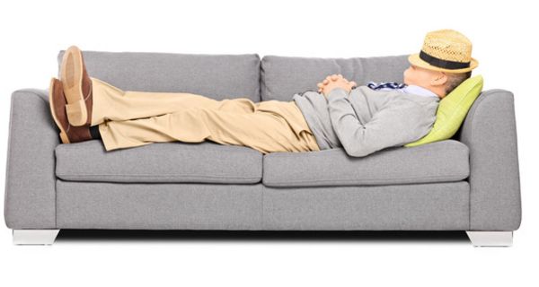 Seitenansicht: Mann um die 60, Strohhut über das Gesicht gelegt, Hände vorm Bauch gefaltet, liegt entspannt auf einer grauen Couch.