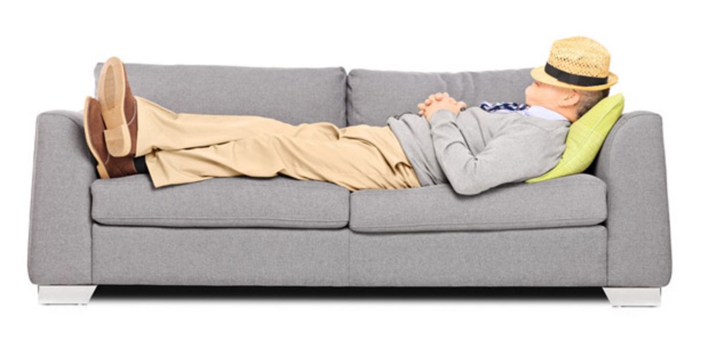 Seitenansicht: Mann um die 60, Strohhut über das Gesicht gelegt, Hände vorm Bauch gefaltet, liegt entspannt auf einer grauen Couch.