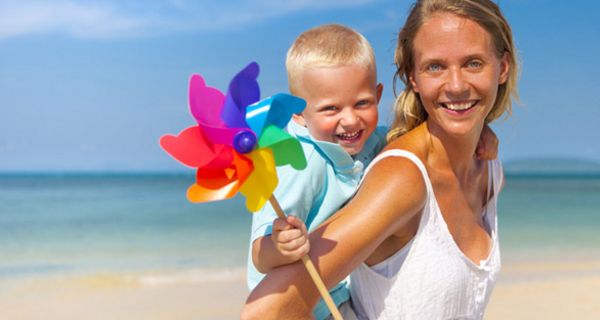 Heiteres Strandfoto: Blonde, lachende Frau, bis Hüfte zu sehen, weißes Shirt, in die Kamera lachend, auf dem Rücken ein kleiner Junge mit blonden Haaren, ein buntes Windrad in der rechten Hand