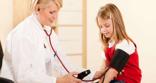 Ärztin misst Blutdruck bei einem Mädchen, das im roten T-Shirt neben ihr sitzt.