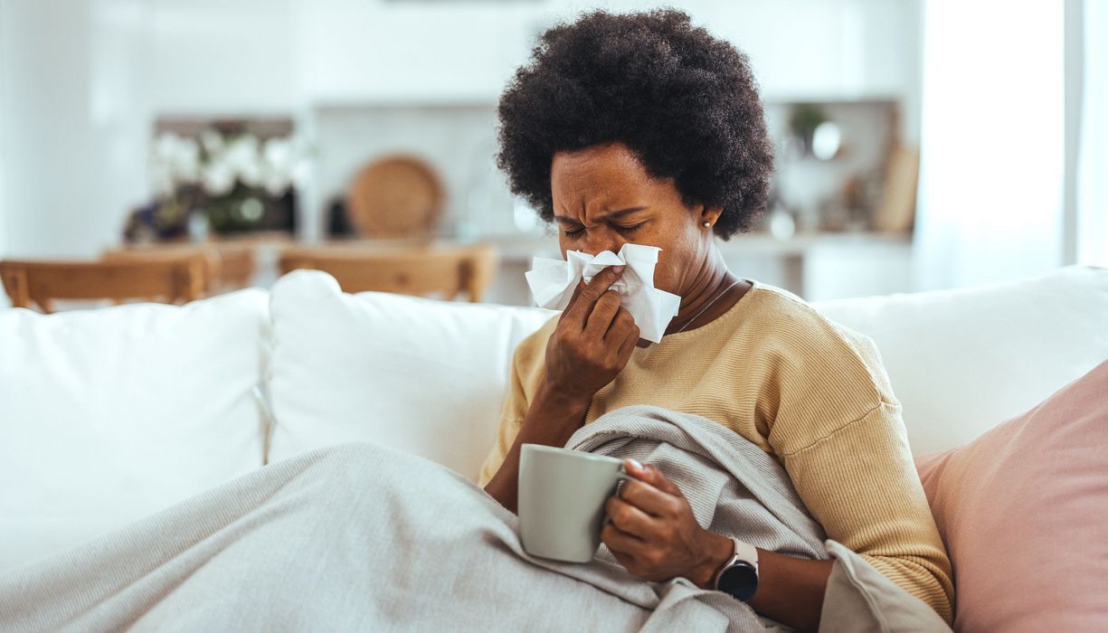 Eine Frau sitzt zugedeckt auf dem Sofa und schnäutzt sich die Nase. Sie hält eine Tasse in der Hand.
