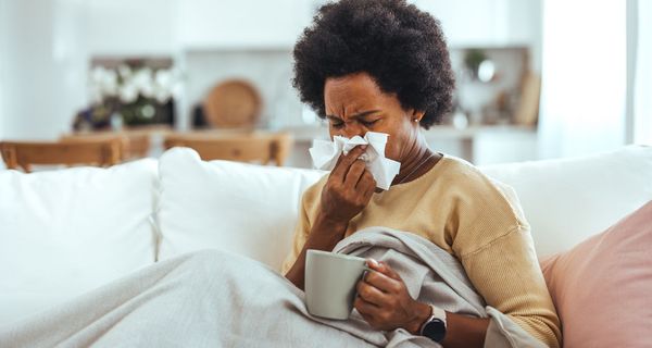 Eine Frau sitzt zugedeckt auf dem Sofa und schnäutzt sich die Nase. Sie hält eine Tasse in der Hand.