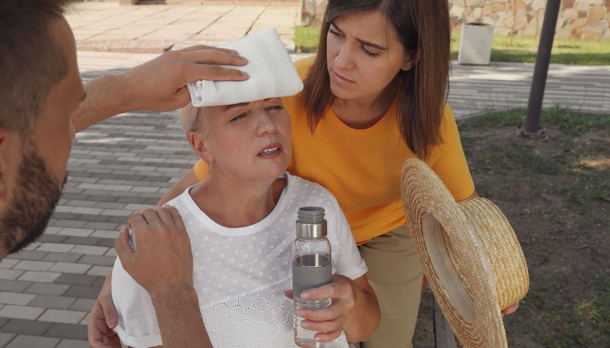 Frau mit Hitzschlag mit zwei Helfern im Schatten: Ein Helfer hält der Frau einen Eisbeutel an die Stirn. Sie hat bereits eine Wasserflasche in der Hand, die Helferin stützt die Frau und fächelt ihr Luft zu.