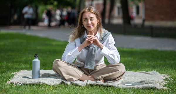 Junge Frau sitzt im Business-Outfit auf einer kleinen Decke im Gras in einem Stadtpark, Fußweg und Gebäude im Hintergrund. 