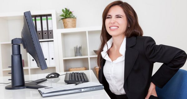 Frau im Büro am Schreibtisch mit PC hält sich den schmerzenden unteren Rücken