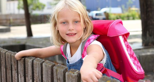 Mädchen, ca. 8 Jahre alt, blond, mit rosafarbenem Schulranzen, über einen Zaun gelehnt und sich dran festhaltend, schaut in die Kamera