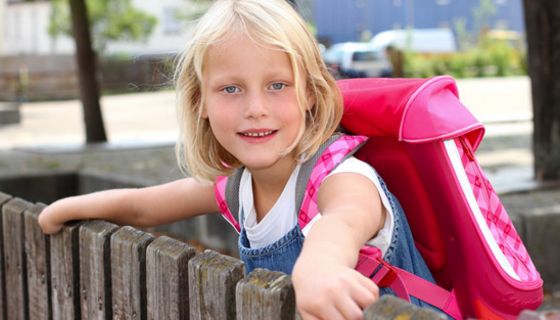 Mädchen, ca. 8 Jahre alt, blond, mit rosafarbenem Schulranzen, über einen Zaun gelehnt und sich dran festhaltend, schaut in die Kamera