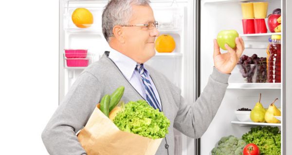 Senior, ca. Ende 60, in der Hand einen Apfel haltend, mit Papiertüte voller Gemüse vor geöffnetem Kühlschrank, darin Salat, Tomaten, Getränke etc.