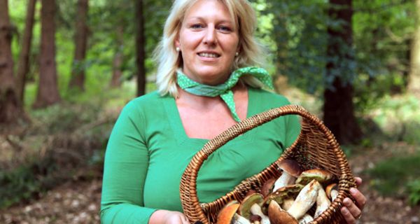 Frontalfoto, Frau in den 40ern, blond, türkises Langarmshirt, türkises Nickituch, im Wald, Korb mit Pilzen in den Händen