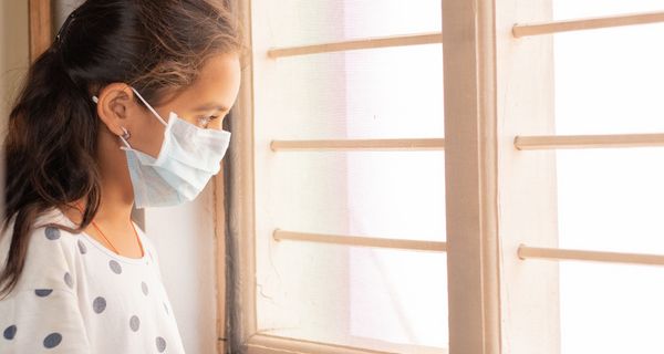 Junges Mädchen mit Maske, steht am Fenster und schaut nach draußen.