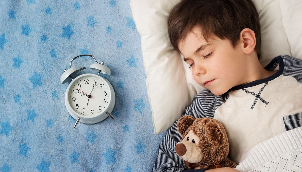 Ein Junge schläft mit einem Teddy neben einem Wecker.