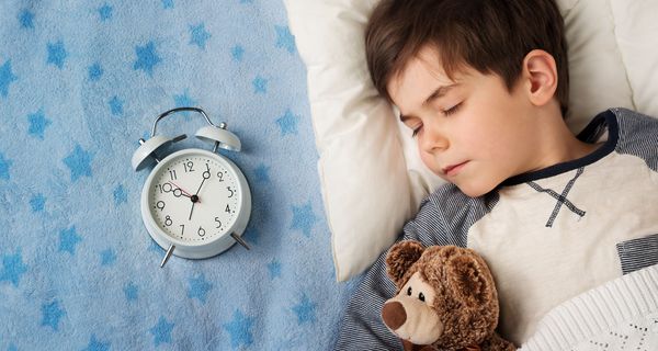 Ein Junge schläft mit einem Teddy neben einem Wecker.