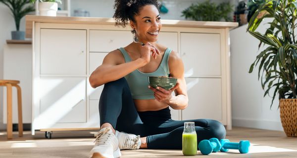 Junge Frau in Sportkleidung, neben sich Hanteln, sitzt auf dem Küchenboden, isst einen Salat.