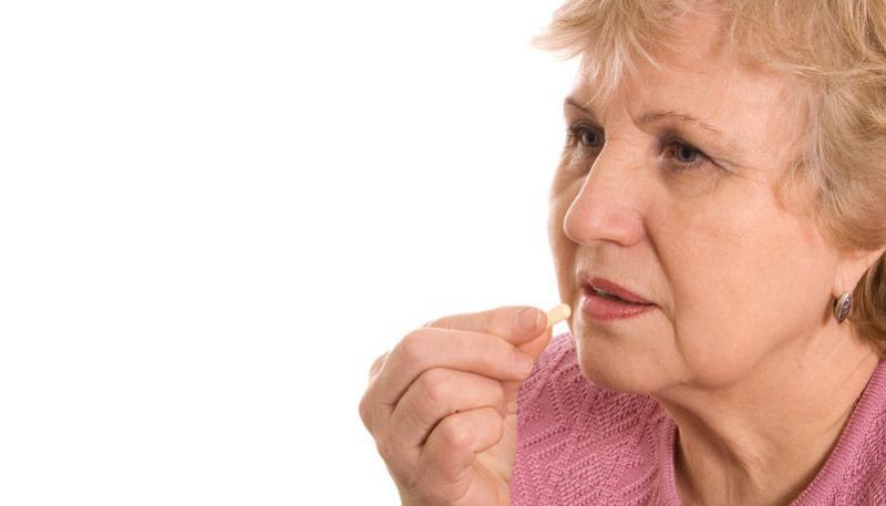 Ältere Frau in rosa Strickjacke führt Tablette mit recher Hand zum Mund, Glas Wasser in der anderen Hand