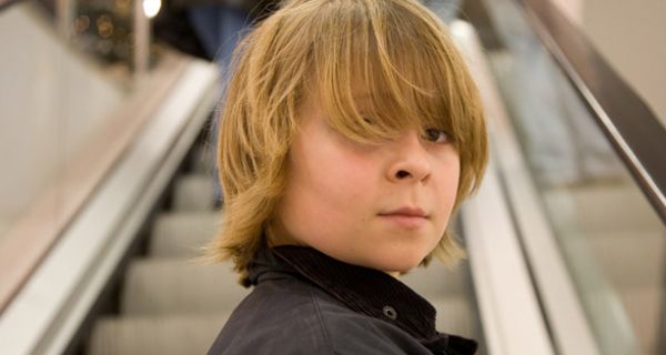 Junge, ca. 12 Jahre, blond, langer Pony, auf der Rolltreppe eines Shopping-Centers, über Schulter in die Kamera blickend