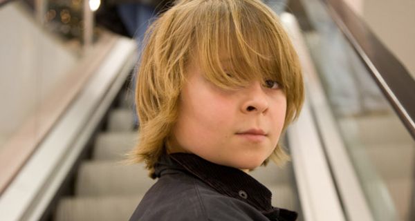 Junge, ca. 12 Jahre, blond, langer Pony, auf der Rolltreppe eines Shopping-Centers, über Schulter in die Kamera blickend
