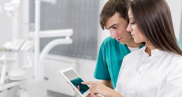 Junge Ärztin und Arzt im Profil am rechten Bildrand mit Tablet (Ärztin); im Hintergrund Zahnarztstuhl