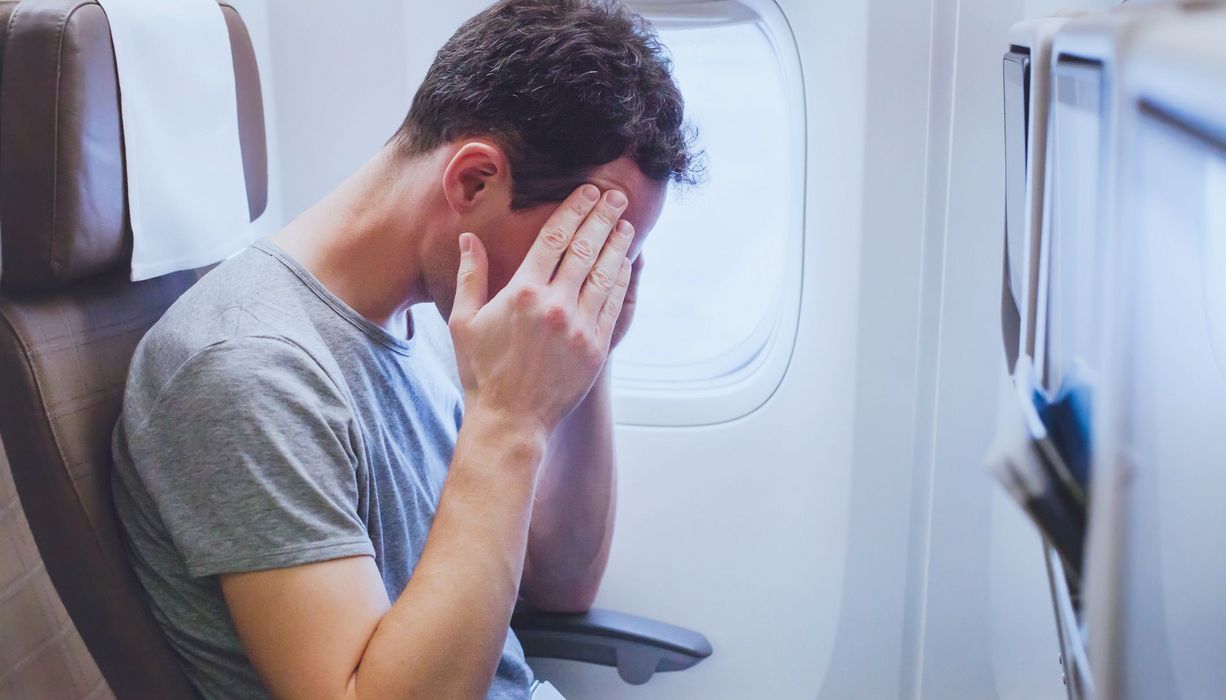 Junger Mann, sitzt in einem Flugzeug und hat starke Kopfschmerzen.