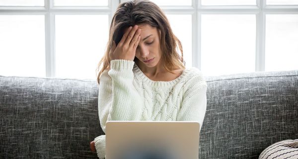 Frau mit Laptop auf dem Schoß, fasst sich an die Schläfe.