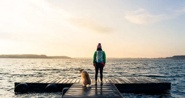 Junge Frau, mit dem Rücken zur Kamera, neben ihr ein Hund, beide stehen an einem Steg mit Blick auf das Wasser.