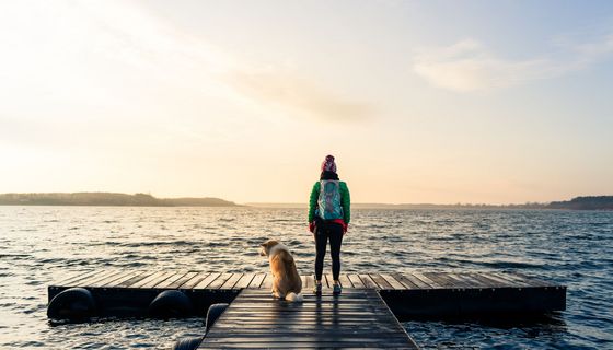 Junge Frau, mit dem Rücken zur Kamera, neben ihr ein Hund, beide stehen an einem Steg mit Blick auf das Wasser.