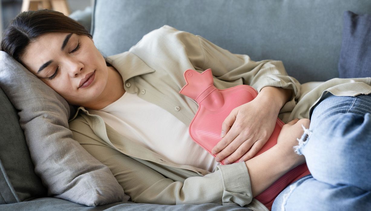 Junge Frau ruht sich auf einem Sofa aus, eine Wärmeflasche auf dem Bauch.