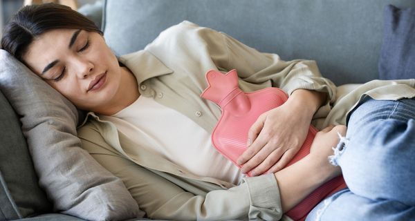 Junge Frau ruht sich auf einem Sofa aus, eine Wärmeflasche auf dem Bauch.