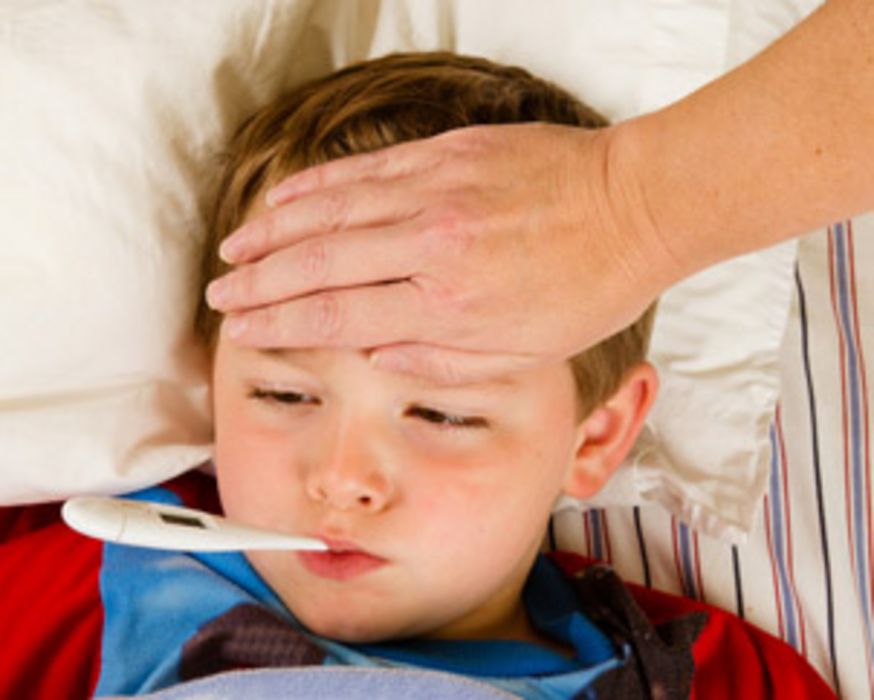 Junge im Kindergartenalter liegt mit Fieberthermometer im Mund im Bett und die Hand eines Erwachsenen liegt auf seiner Stirn.
