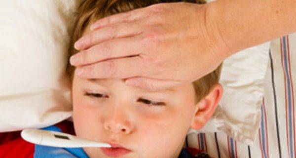 Junge im Kindergartenalter liegt mit Fieberthermometer im Mund im Bett und die Hand eines Erwachsenen liegt auf seiner Stirn.