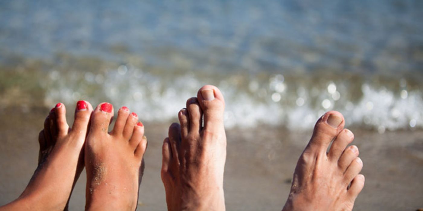 Je ein Paar Frauen- und Männerfüße strecken sich am unteren Bildrand ins Bild, dahinter Strand und Meer