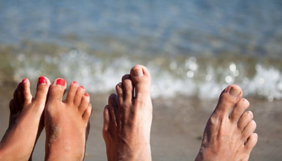 Je ein Paar Frauen- und Männerfüße strecken sich am unteren Bildrand ins Bild, dahinter Strand und Meer