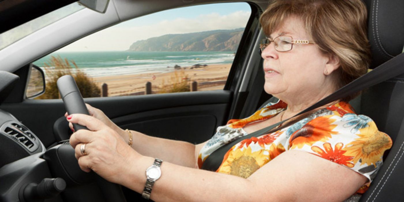 Seniorin im bunten Sommerkleid im linken Profil hinterm Steuer ihres Autos, im Hintergrund mediterrane Küstenlandschaft und Meer