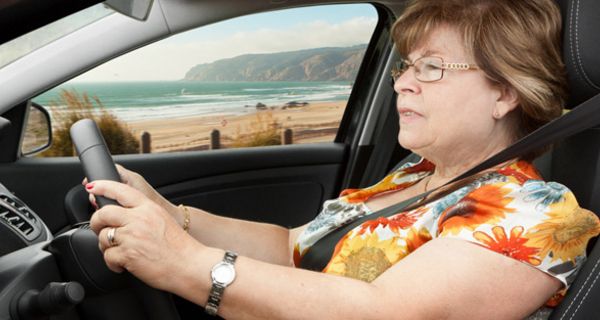 Seniorin im bunten Sommerkleid im linken Profil hinterm Steuer ihres Autos, im Hintergrund mediterrane Küstenlandschaft und Meer