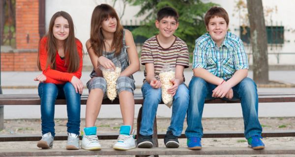 Zwei Mädchen und zwei Jungen, junges Teenageralter, sitzen auf der Lehne einer Holzbank, Füße auf der Bank; der Junge rechts ist übergewichtig